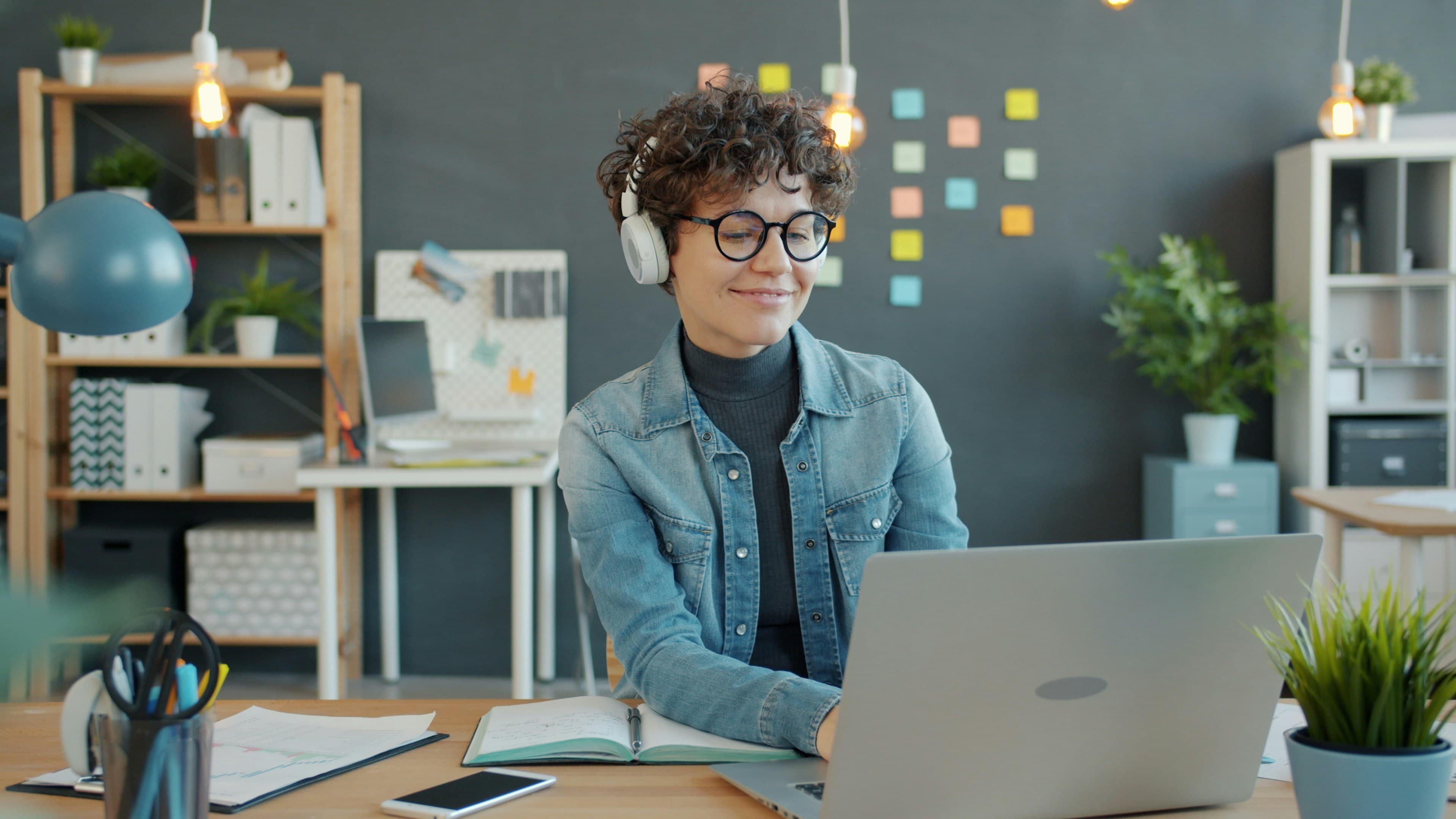 A woman in headphones working on a laptop at an office desk